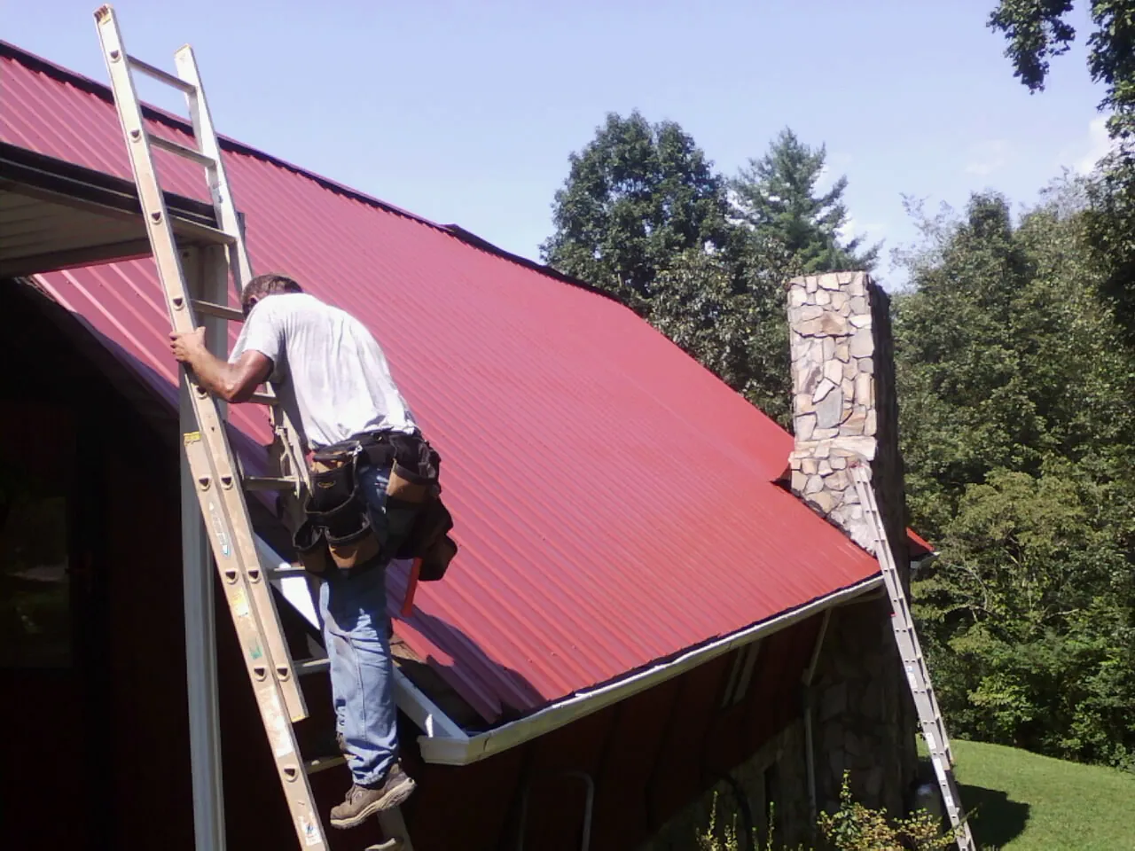 Roofer on a ladder beside a red metal roof and stone chimney — Soffit & Fascia Repair by Roofing Brothers in Jefferson Valley